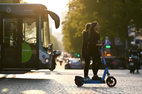 Deux jeunes femmes attendent sur une trottinette électrique, baignées par la lumière du soleil couchant. Un bus RATP est garé à gauche.