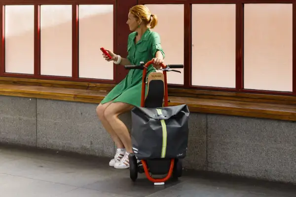 Femme en robe verte assise, tenant un téléphone rouge, à côté d'une trolley trottinette grise et orange.