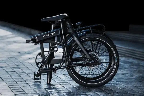 Un vélo électrique carbone pliant, noir, sur un pavé mouillé. Son design moderne et épuré promet des trajets urbains fluides.