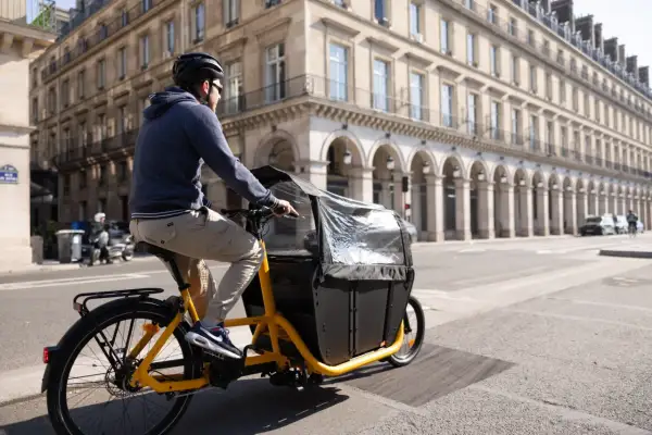 Un homme roule à Paris sur un vélo électrique cargo, prêt à transporter une charge lourde dans sa caisse noire.