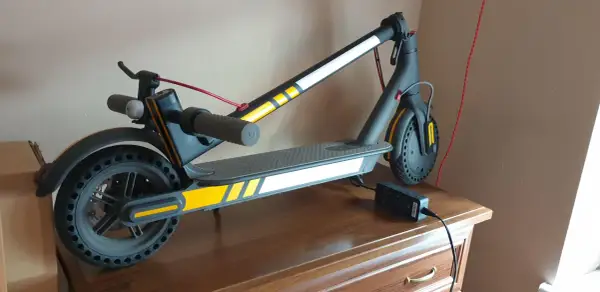 Trottinette électrique noire avec des bandes jaunes et blanches, des stickers réfléchissants pour plus de sécurité. Elle est branchée à son chargeur.