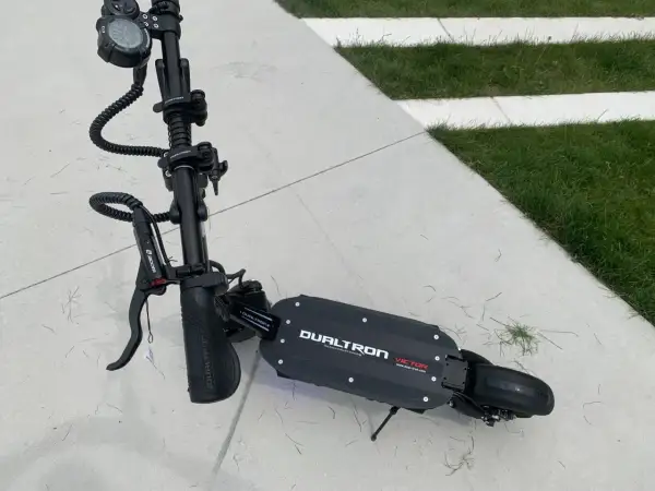 Trottinette électrique Dualtron Victor, noire, sur un sol en dalles de béton et herbe. Le poids de ce modèle est un atout pour sa maniabilité.