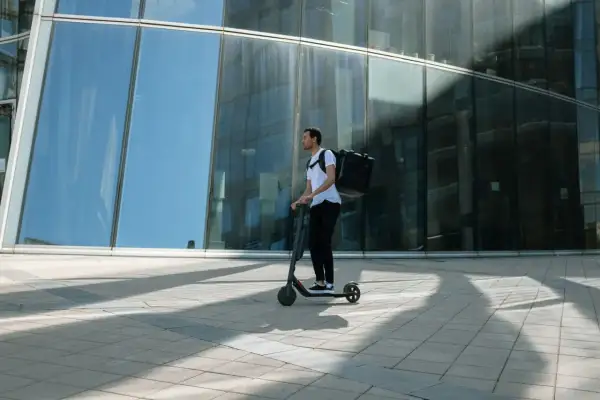Livreur sur trottinette avec une grande sacoche noire, se déplaçant devant un immeuble moderne aux vitres bleues.