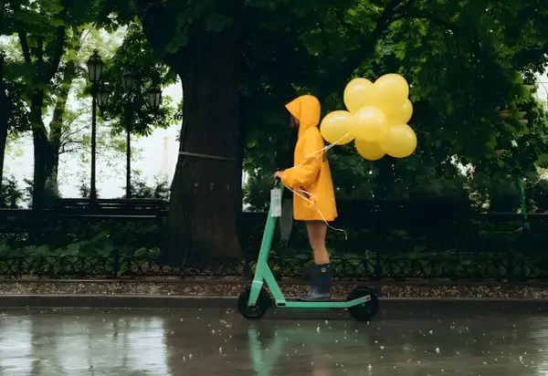 Une personne en manteau jaune conduit une trottinette ipx5 sous la pluie, tenant des ballons jaunes.